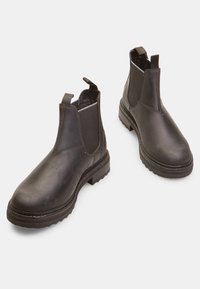 Botas de tobillo de piel negra con paneles laterales elásticos, lengüetas en el bucle y suelas de goma texturizadas, con una superficie suave y un diseño de puntera redondeada.