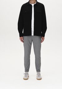 Man wearing black buttoned shirt over white t-shirt, gray checkered pants, and white sneakers standing against a plain background.