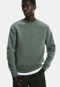 Zielony sweter z miękkiego materiału z okrągłym dekoltem. Posiada małe logo na klatce piersiowej oraz swobodny krój. Nałożony na biały T-shirt.