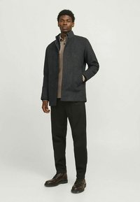 Dark gray wool blend coat with a stand collar, button closure, two side pockets, paired with brown pants and boots. Smooth texture.