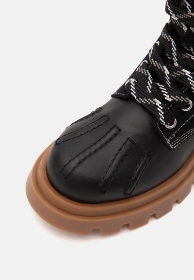 Botte en cuir noir avec semelle épaisse en caoutchouc marron, couture décorative sur la pointe et lacets à motifs noirs et blancs sur fond blanc.