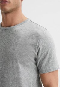 Tricou gri dintr-un material moale, cu guler rotund și mâneci scurte, având o suprafață ușor texturată și detalii de coasere minime.