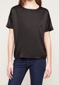 Top corto de satén negro con cuello redondo, que presenta un corte relajado y una textura suave. Combinado con jeans de color azul oscuro.