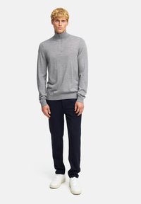 Grauer Half-Zip-Pullover aus leichtem Material, runder Halsausschnitt, gerippte Bündchen, kombiniert mit dunkelblauen Hosen und weißen Sneakers.