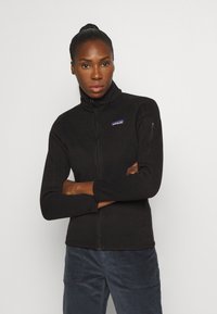 Zwarte fleece jack met ritssluiting en hoge kraag, met een klein paars Patagonia-logo op de borst en een zijzak met rits. Zachte textuur.