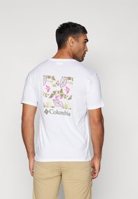Hvit kortermet t-skjorte med blomsterdesign i rosa og grønt på baksiden, med Columbia-logoen i grått under designet.