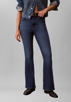 Sötétkék bootcut farmernadrágot, fekete négyzetormú cipőt és felhajtott farmerdzsekit viselő személy, kéz állítja a fekete övcsatot.