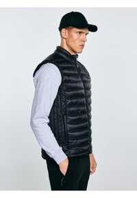 Fiatal férfi fekete sapkában, fekete steppelt mellényben és szürke pulóverben, profilból állva, kezét zsebeiben tartva, sima világos háttér előtt.