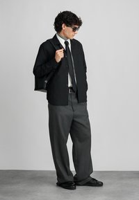 Camisa preta de botões sobre uma camisa branca, calças cinzentas e sapatos pretos. Apresenta um bolso no peito e uma mala tote preta. Óculos de sol escuros usados.