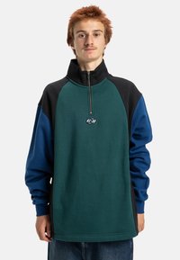 Pulôver de fleece em cores bloqueadas, apresentando um colarinho com zíper de metade, com painéis verdes, pretos e azuis. Inclui um detalhe de logótipo no peito.