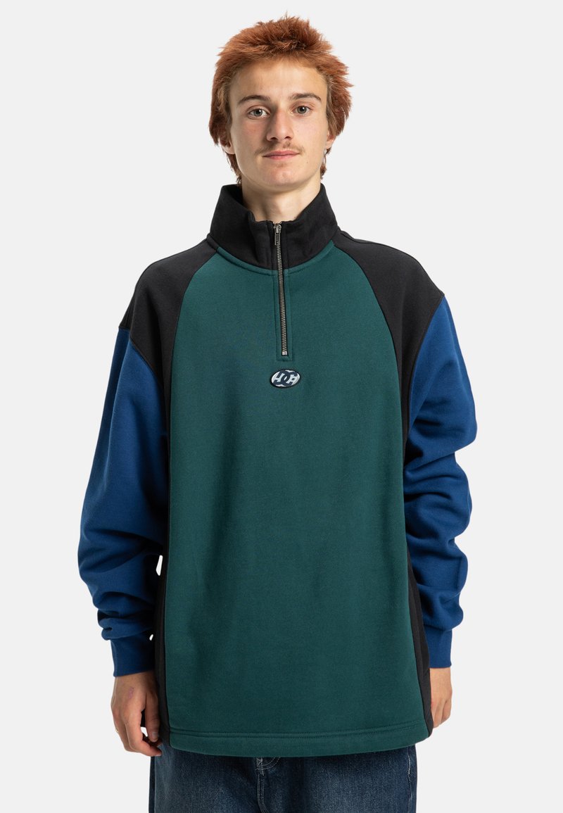 Pulôver de fleece em cores bloqueadas, apresentando um colarinho com zíper de metade, com painéis verdes, pretos e azuis. Inclui um detalhe de logótipo no peito.