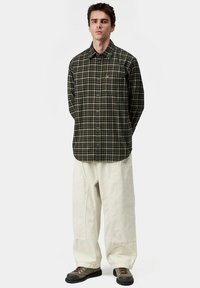 Camisa de cuadros en verde oscuro y beige, combinada con pantalones sueltos beige y calzado oscuro. Cuenta con un cuello con solapa y un bolsillo en el pecho.