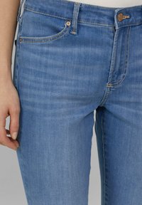 Gros plan sur une personne portant un jean en denim bleu avec des coutures visibles, des poches avant et une fermeture à bouton, la main reposant à côté de la cuisse.