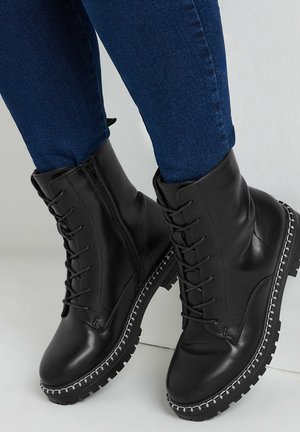 Stivaletti neri in pelle con chiusura a lacci e cerniera, suola robusta, dettagli in cucitura argentata e texture liscia, abbinati a jeans blu scuro.