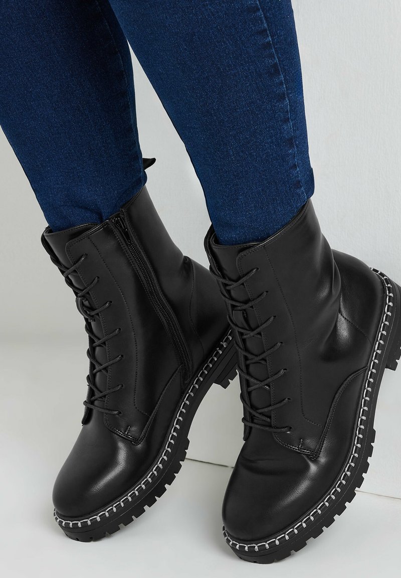 Botas de tobillo de cuero negro con cordones y cremallera, suela robusta, detalles de costura plateada y textura suave, combinadas con unos vaqueros azul oscuro.