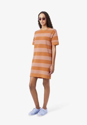 Mujer con vestido de rayas cortas naranja y rosa, gafas de sol rosas, zapatillas azules y una pulsera, de pie frente a un fondo blanco liso.