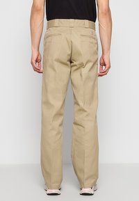 Pantalon beige à jambes droites avec poches arrière porté par une personne en chemise noire et baskets blanches, debout devant un fond clair uni.