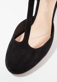 Musta mokkanahkasaapas, jossa on mantelinmuotoinen kärki ja leikattu yksityiskohta sivuilla. Varustettu sileällä beigen värisellä sisäpohjalla ja minimalistisella muotoilulla.