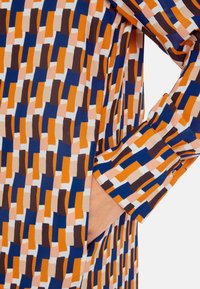 Bunte Bluse mit einem geometrischen Muster in Orange, Blau, Creme und Braun, mit langen Ärmeln und einem geknöpften Bündchen. Enthält eine Seitentasche.