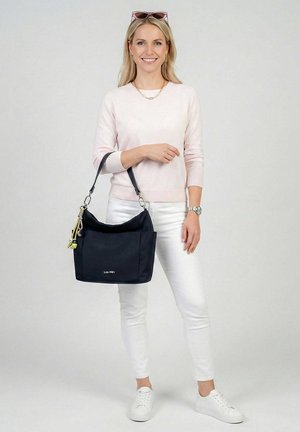 Donna con maglione rosa chiaro e pantaloni bianchi che tiene una borsa nera a tracolla, indossa scarpe da ginnastica bianche e occhiali da sole sulla testa, sorridente.