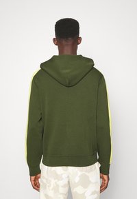 Olivgrön hoodie med huva, med gula sidosträck. Tyget är slätt med ribbade muddar och fåll, visad bakifrån.