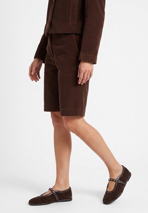 Person iført brune kordfløjls-shorts, matchende jakke og mørkebrune flade sko med ankelremme, går på en hvid baggrund.