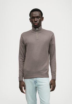 Uomo che indossa un pullover taupe a maniche lunghe con zip al quarto e jeans azzurri chiari, in piedi davanti a uno sfondo bianco uniforme.