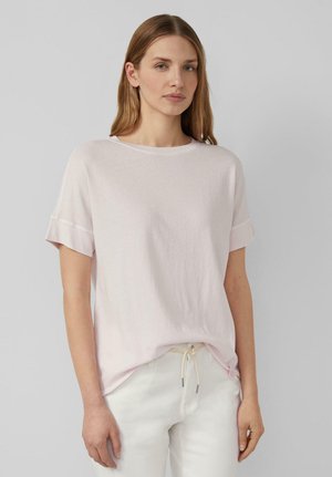 Frau mit glattem hellbraunem Haar trägt ein blassrosa kurzärmliges Oberteil und weiße Hose mit Kordelzug vor einem schlichten grauen Hintergrund.