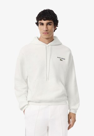 Jongeman met donker krullend haar, gekleed in een witte hoodie met een voorzak en de tekst "Jeu Set et Match" en een klein groen logo op de borst.