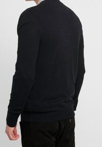 Pull marin sombre avec un col rond, des poignets et un ourlet côtelés, et une texture en maille lisse. Porté sur un pantalon noir.