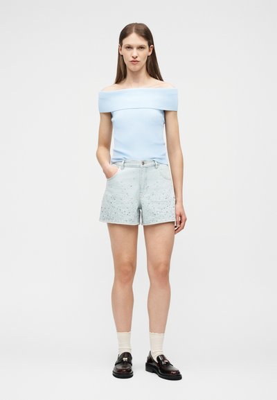 Γυναίκα που φοράει ανοιχτό μπλε τοπ χωρίς ώμους, σκισμένες τζιν σορτς με στίγματα, κρεμ κάλτσες και μαύρα loafers, στέκεται με το ένα χέρι στην τσέπη.