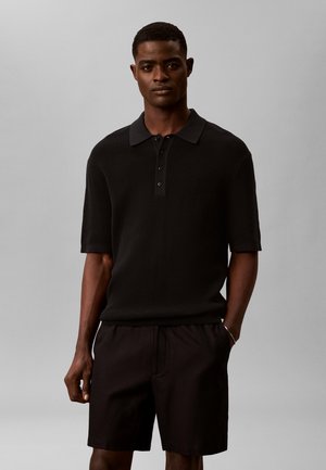 Άνδρας στέκεται με το ένα χέρι στην τσέπη φορώντας μαύρο πλεκτό πόλο με κοντά μανίκια και μαύρα σορτς μπροστά σε απλό φόντο.