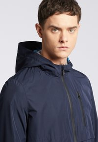 Granatowa kurtka z kapturem, wykonana z lekkiego, wodoodpornego materiału. Posiada zamek błyskawiczny z przodu oraz kieszeń na piersi o eleganckim, minimalistycznym designie.