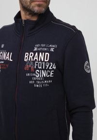 Felpa con zip di colore blu navy realizzata in tessuto morbido, con grafiche testuali bianche e rosse, tasche laterali e dettagli di cucitura a contrasto.