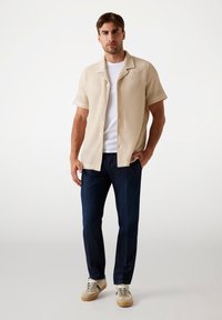 Short-sleeve, beige striped button-up shirt over a white t-shirt, paired with dark blue jeans and beige sneakers with green accents.