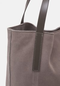 Szary zamszowy tote bag o miękkiej teksturze, z ciemnoszarymi skórzanymi uchwytami i widocznymi szwami wzdłuż szwów. Projekt otwarty na górze.