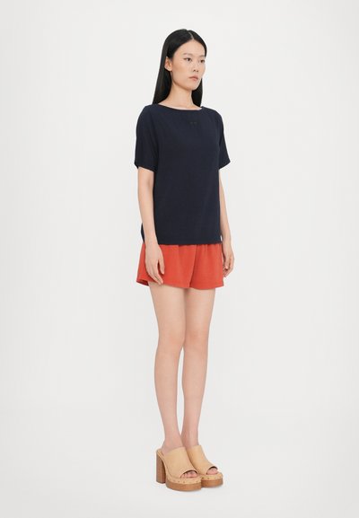 Top corto blu navy a maniche corte con scollatura a barca e una sottile texture, abbinato a pantaloni rossi. Il modello indossa zoccoli color sabbia con base in legno.