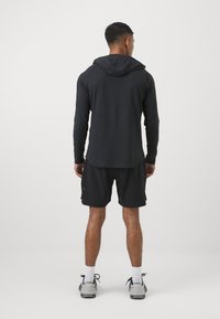 Schwarzes Langarm-Hoodie-Shirt kombiniert mit schwarzen Shorts. Das Material scheint leicht zu sein. Sneakers mit grauen und schwarzen Akzenten.
