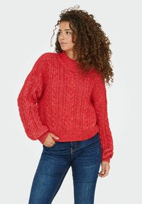 Rode kabelgebreide trui met een hoge, geribbelde halslijn en volumineuze mouwen, gecombineerd met donkerblauwe jeans en een gestructureerd breipatroon.