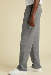 Vista lateral de una persona que lleva pantalones de chándal grises y zapatillas blancas, de pie contra un fondo beige liso.