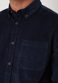 Mørkeblå kordfløjlsskjorte med knap-ned krave, der har en brystlomme og tekstureret stof. Kendt for sit ribbede design og knapper.