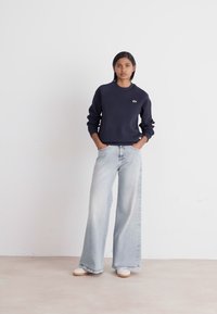 Marineblå sweatshirt med rund hals og lille logo, parret med lyseblå brede jeans. Model iført hvide sneakers på en ensfarvet baggrund.