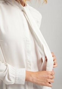 Bílá blůza s límečkem a knoflíkovou légou, vybavená dlouhými rukávy s nařasenými manžetami a hladkou texturou.