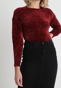 Burgunderfarbener Strickpullover mit langen Ärmeln und rundem Ausschnitt, kombiniert mit einem hoch geschnittenen schwarzen Jeansrock. Glatte Textur und figurbetonter Schnitt.