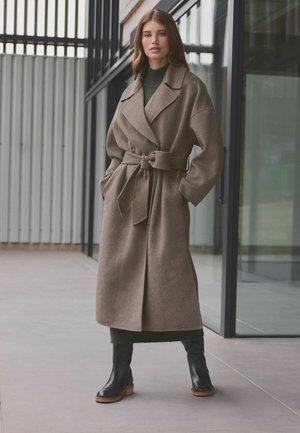 Cappotto in misto lana beige con design avvolgente, grandi rever, cintura in vita e due tasche laterali. Abbinato a stivali in pelle neri.