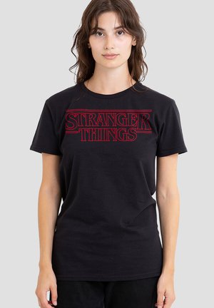 Junge Frau mit braunen Haaren, die ein schwarzes T-Shirt mit rotem "Stranger Things"-Logo vor einem schlichten grauen Hintergrund trägt.