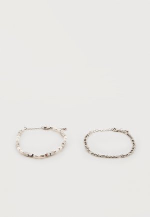 Deux bracelets sur fond blanc : un en perles et chaîne de perles argentées, l'autre une fine chaîne argentée avec un fermoir.