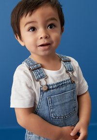 Denim overalls med metalspænder, lomme foran, båret over en hvid t-shirt. Baggrunden er ensfarvet blå. Barnets smilende ansigt.