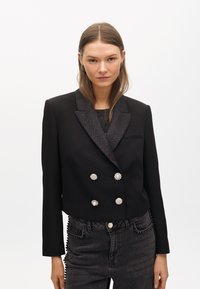 Krój czarnej marynarki z teksturowanym kołnierzem typu szal, podwójnym zapięciem, srebrnymi guzikami oraz eleganckim wykończeniem. Połączona z ciemnymi dżinsami.