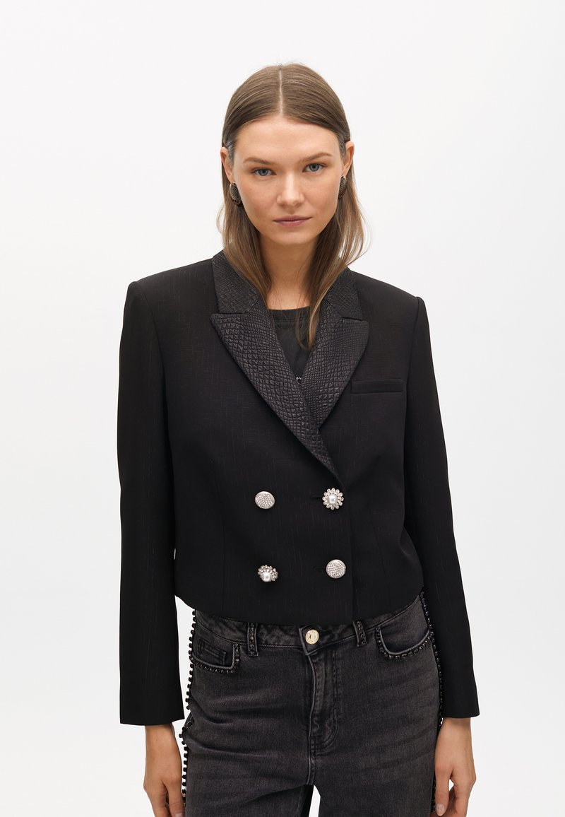 Krój czarnej marynarki z teksturowanym kołnierzem typu szal, podwójnym zapięciem, srebrnymi guzikami oraz eleganckim wykończeniem. Połączona z ciemnymi dżinsami.